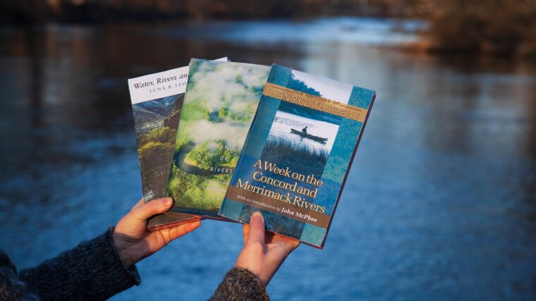 Eine Person hält drei Bücher vor dem Hintergrund eines Flusses