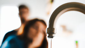 Wasser läuft aus einem Wasserhahn vor zwei schemenhaft erkennbaren Personen im Hintergrund