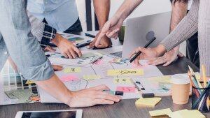Schreibtisch mit Arbeitsmaterialien, Arme von Teammitarbeitern, die auf verschiedene Dinge zeigen