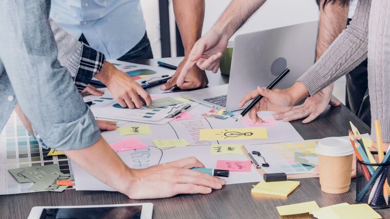 Schreibtisch mit Arbeitsmaterialien, Arme von Teammitarbeitern, die auf verschiedene Dinge zeigen