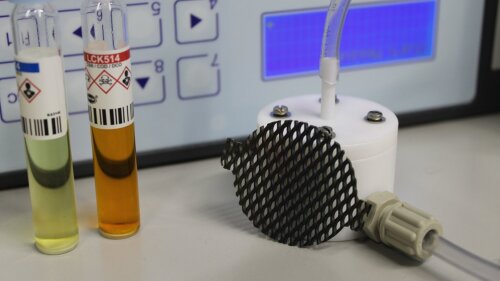 Ein Sensor mit Schlauch-Anschlüssen liegt vor einem elektronischen Gerät und zwei Probenfläschchen mit farbiger Flüssigkeit