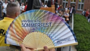 Ein aufgeklappter Fächer mit dem Schriftzug "Universitäts-Sommerfest" vor einer Gruppe von Menschen